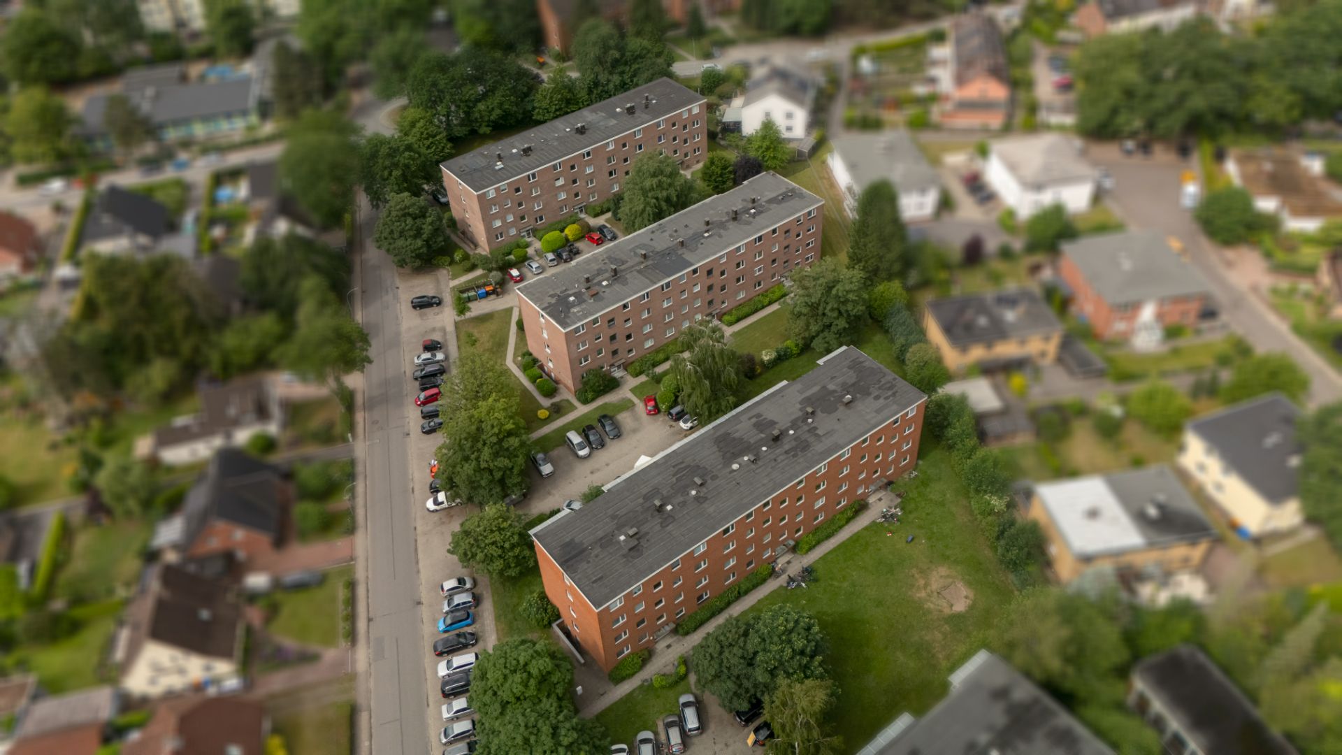 Drei mehrstöckige Wohnhäuser aus rotem Backstein mit Parkplätzen und Bäumen, von oben aufgenommen.