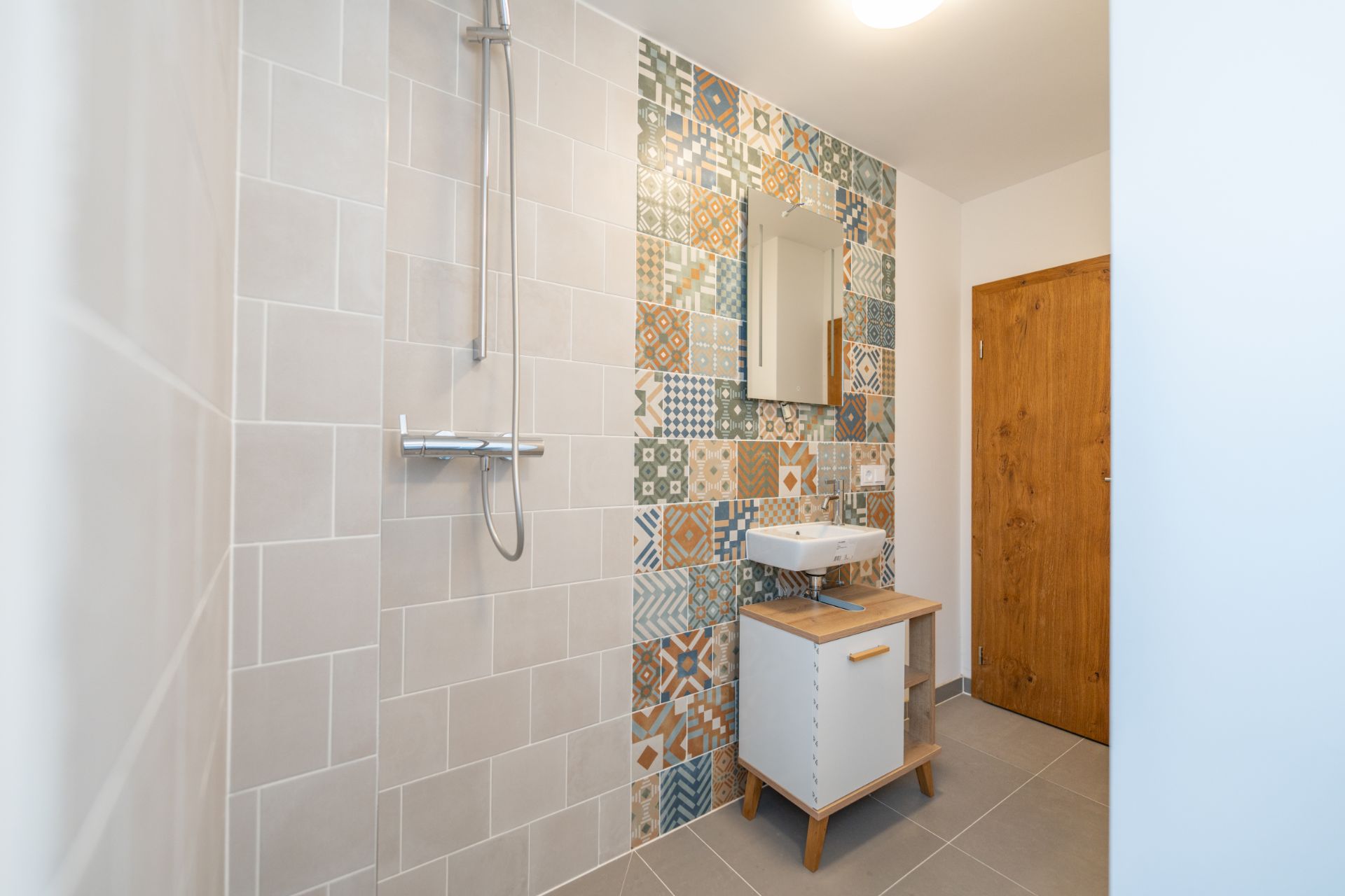 Modernes Badezimmer mit Dusche, bunten Wandfliesen, Waschbecken und Holzschrank.