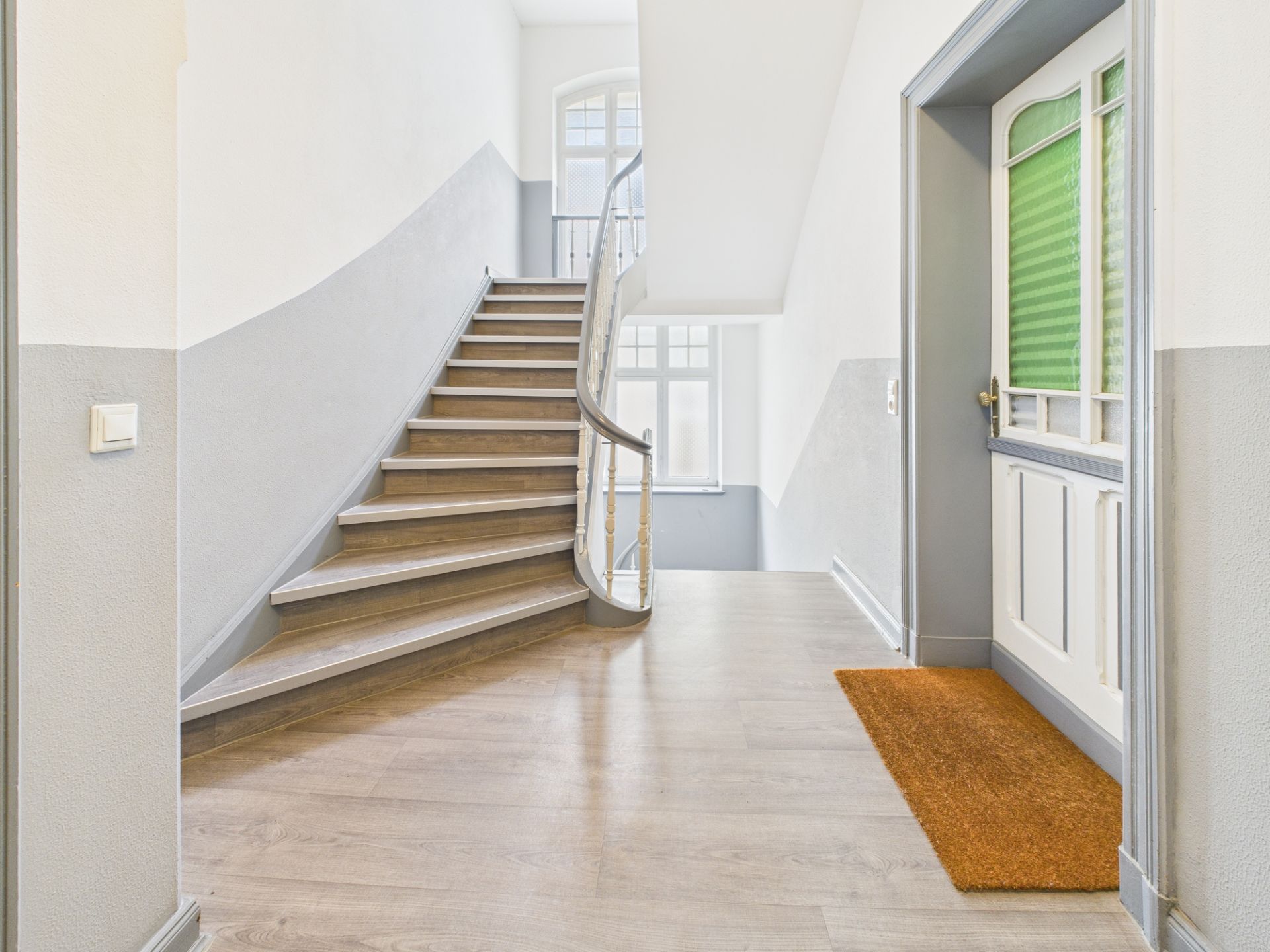 Heller Flur mit Treppe, Tür mit grünem Fenster und orangefarbener Fußmatte.