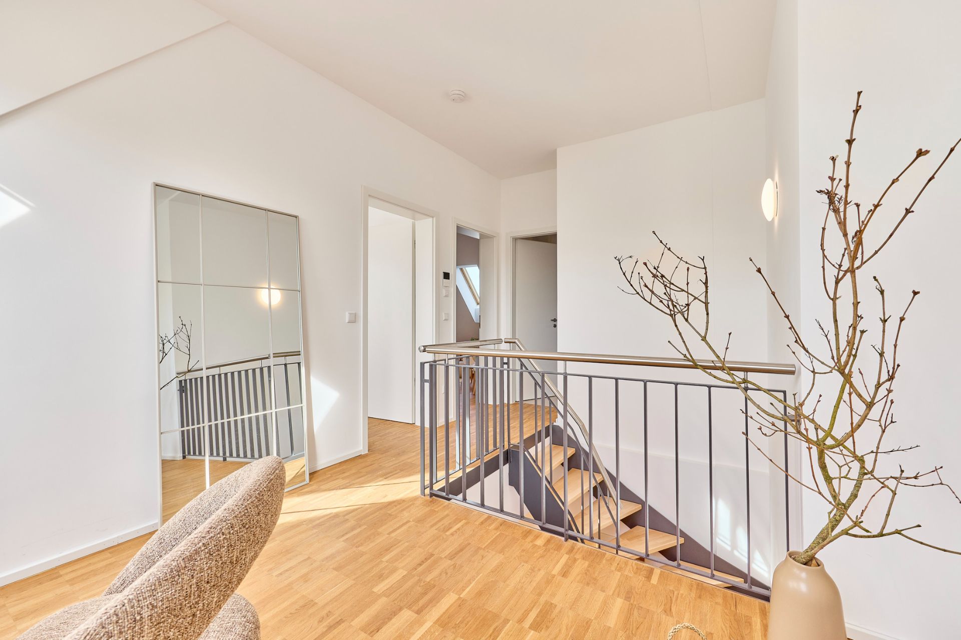 Heller Flur mit Holzfußboden, Treppengeländer, großem Spiegel und dekorativer Vase mit Zweigen.
