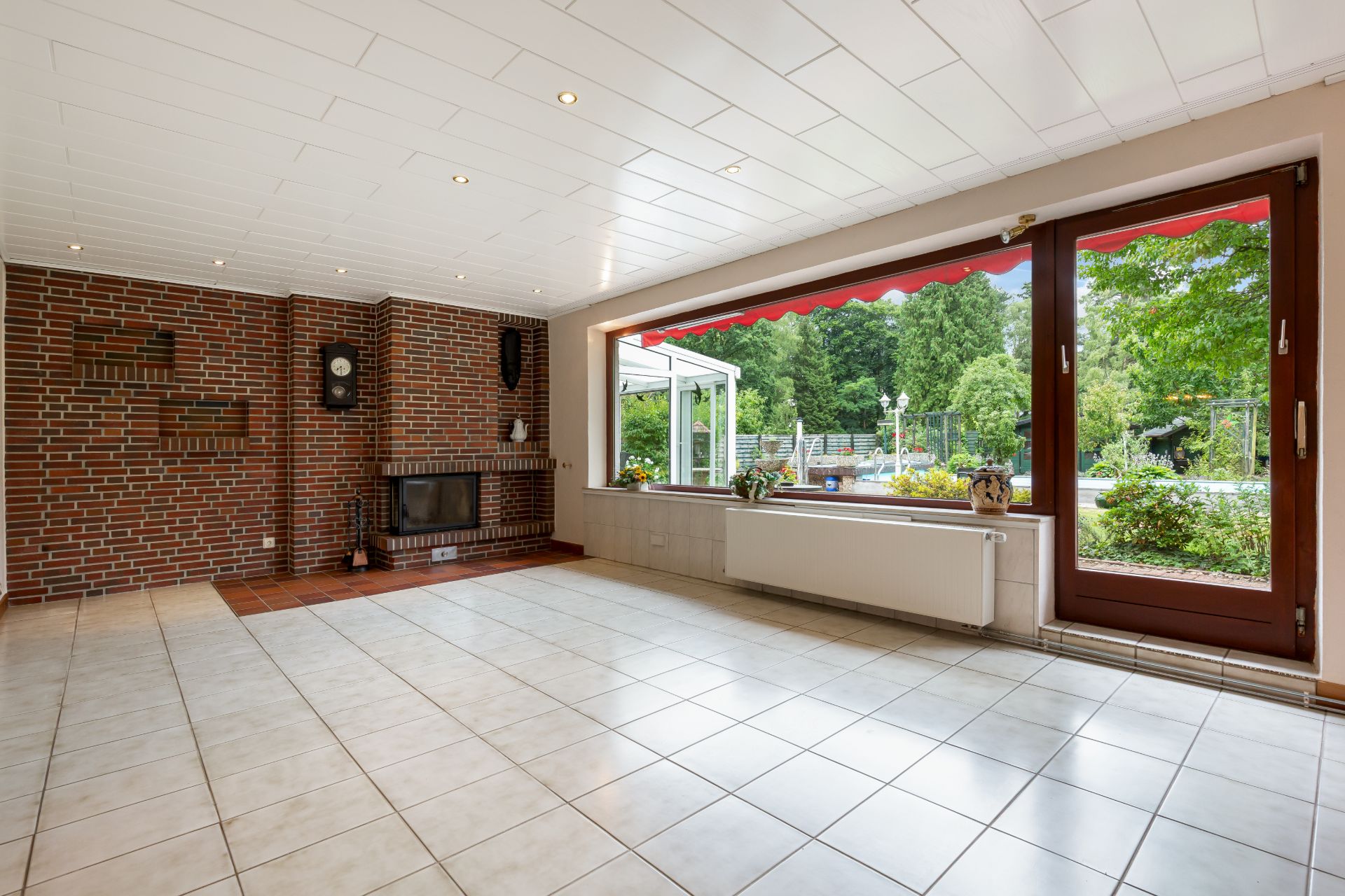 Heller Wohnraum mit Kamin, großen Fenstern und Tür zum Garten, Fliesenboden und Ziegelwand.