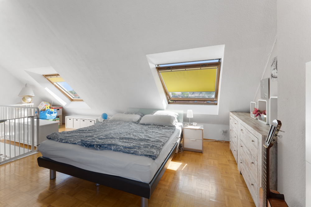Helles Schlafzimmer mit Dachschrägen, Doppelbett, Kommode, Gitterbett und zwei Dachfenstern.