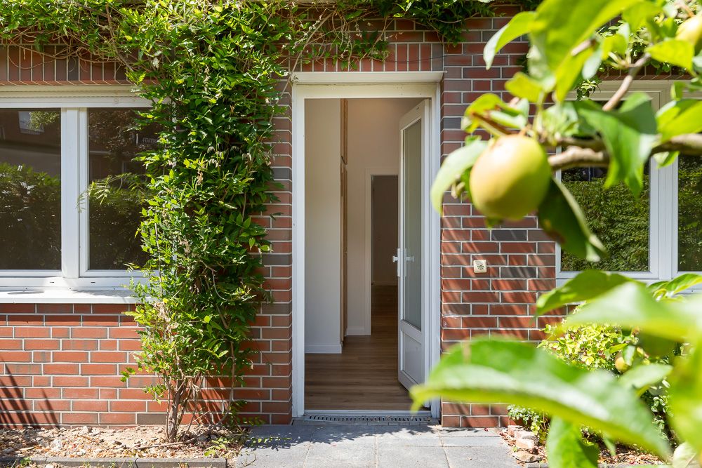Offene Haustür in einer roten Backsteinfassade mit begrüntem Eingang und Apfelbaum im Vordergrund.