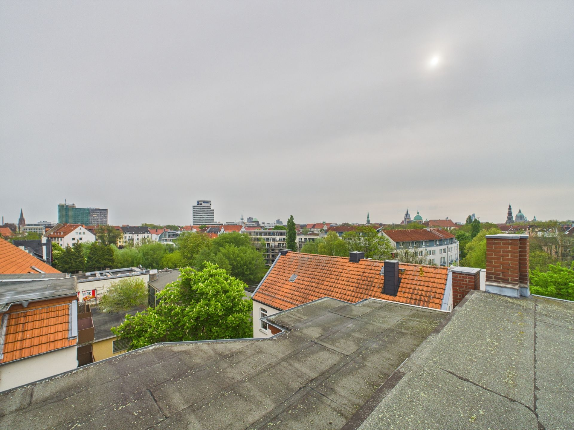 Blick von einem Dach auf rote Ziegeldächer, grüne Bäume und einige Gebäude in der Ferne.