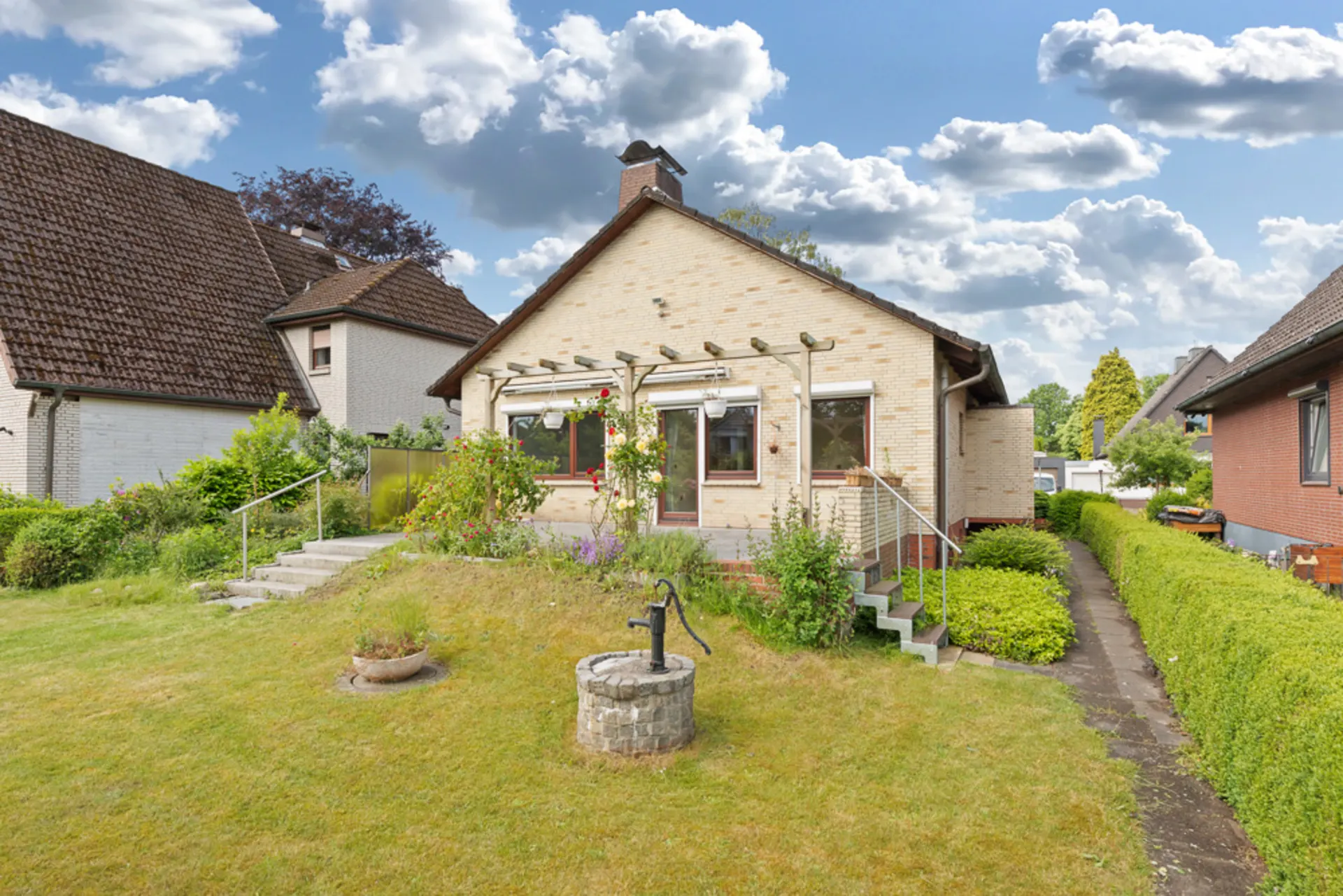 Ein Einfamilienhaus mit hellem Backstein, Garten, Terrasse und einer alten Wasserpumpe im Rasen.