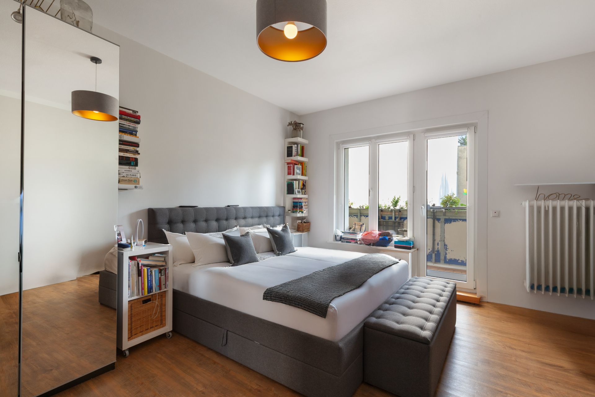 Modernes Schlafzimmer mit Doppelbett, Spiegel, Bücherregalen und großem Fenster mit Balkonzugang.