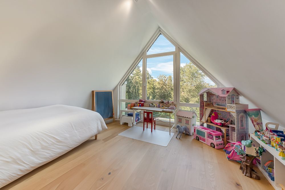 Helles Kinderzimmer mit Spielzeug, Puppenhaus, Bett und großem Fenster mit Blick nach draußen.