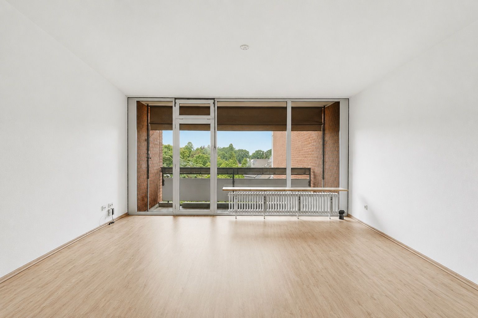 Leeres Zimmer mit Holzfußboden, großen Fenstern und Zugang zu einem Balkon.