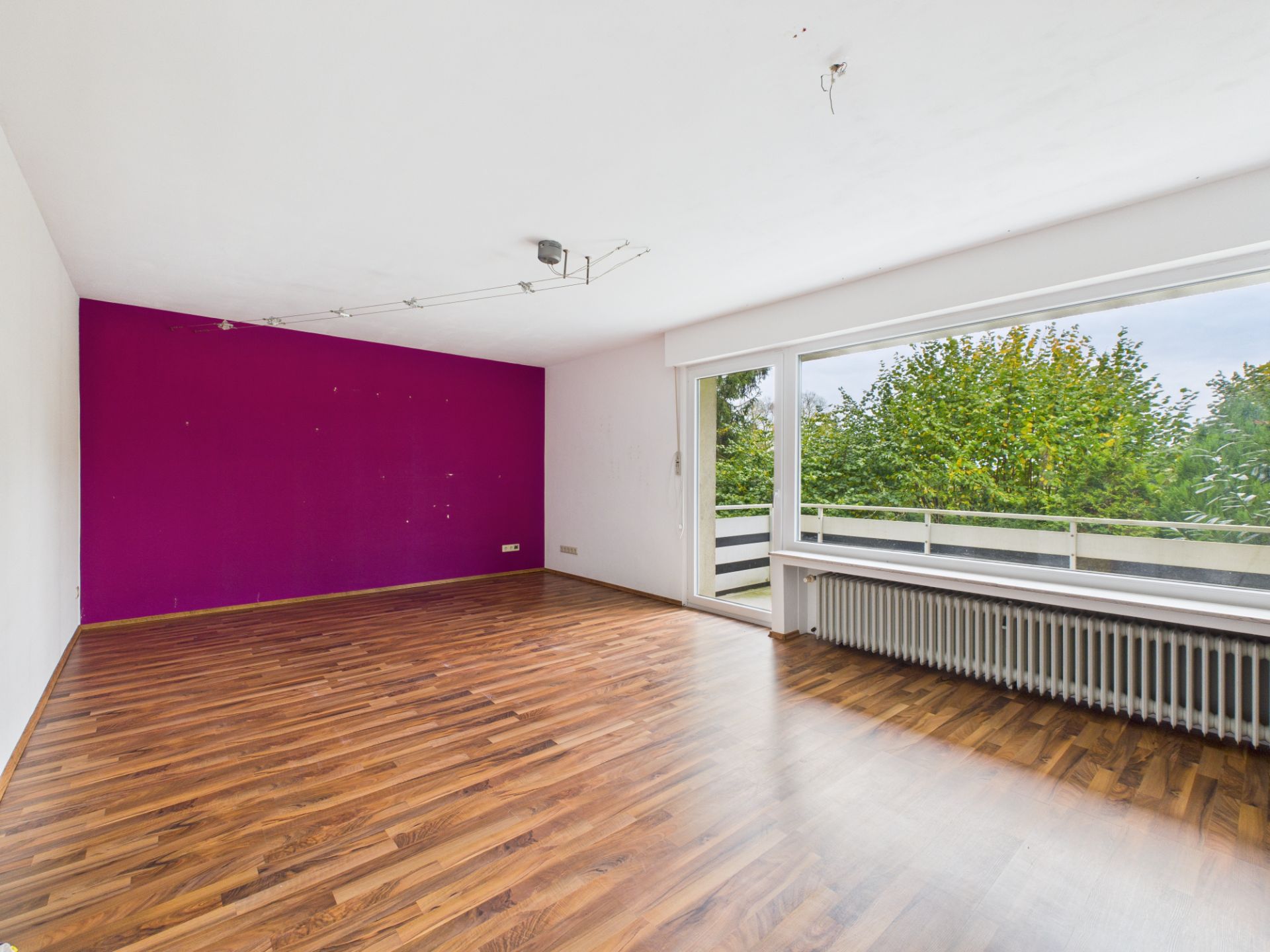 Leeres Zimmer mit lila Wand, Parkettboden und großem Fenster mit Blick ins Grüne.