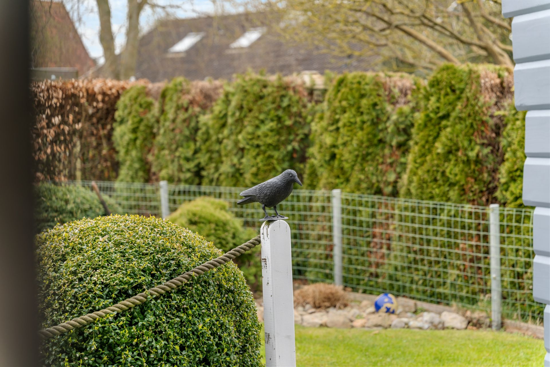 Garten mit Sträuchern, Zaun und einer Vogel-Figur auf einem weißen Pfosten.