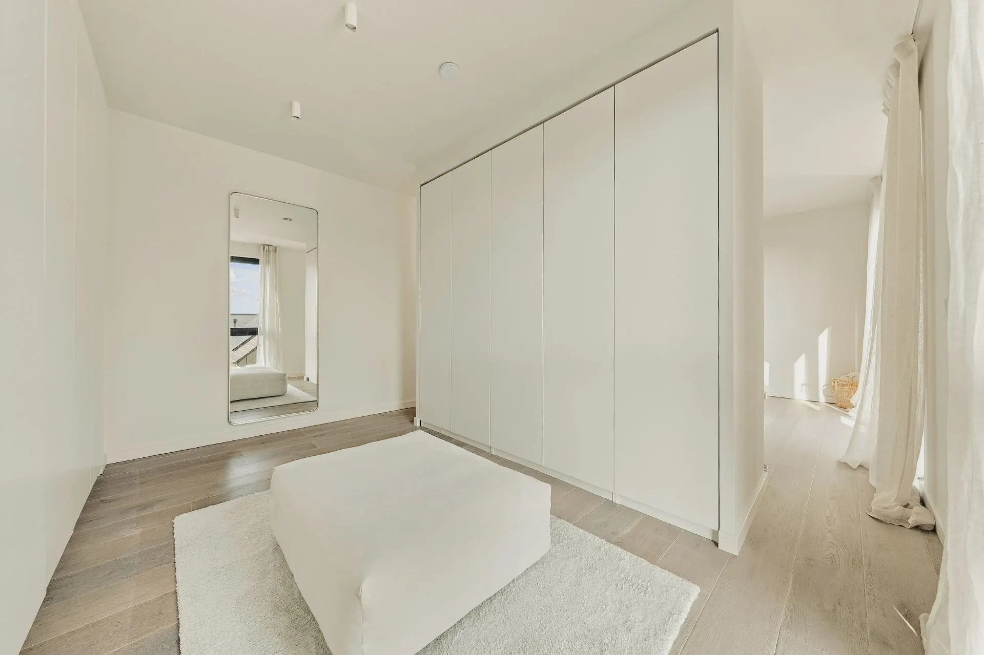 Heller, minimalistischer Ankleideraum mit großem Spiegel, weißem Pouf und Einbauschränken.