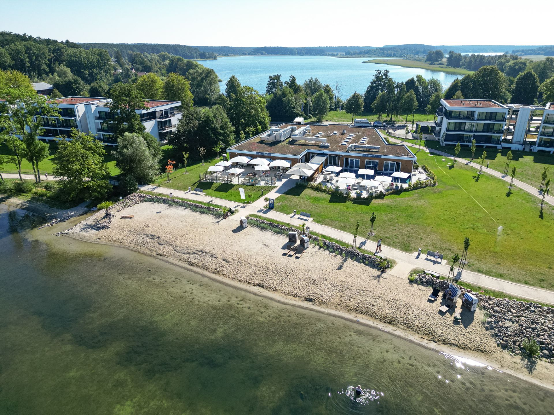 Luftaufnahme von Wohngebäuden mit Garten am Seeufer und kleinem Sandstrand.