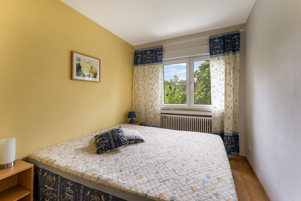 Schlafzimmer mit gelber Wand, Bett, Nachttisch, Fenster mit Ausblick auf Bäume und Vorhängen.