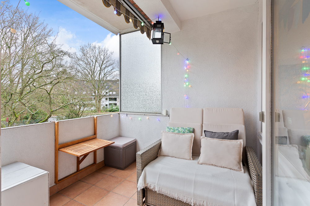 Überdachter Balkon mit Sofa, kleinem Tisch, Sitzhocker und Lichterkette, mit Blick auf Bäume.