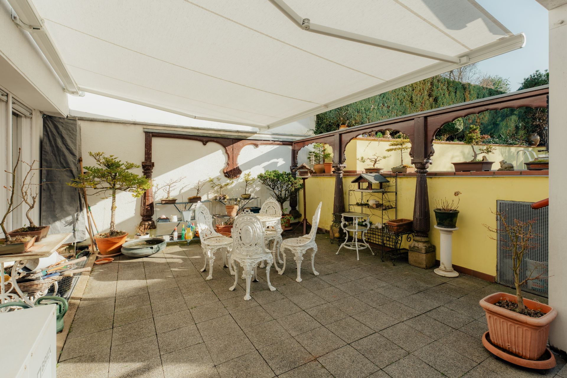 Überdachte Terrasse mit weißen Gartenmöbeln, Pflanzen und gelber Wand.