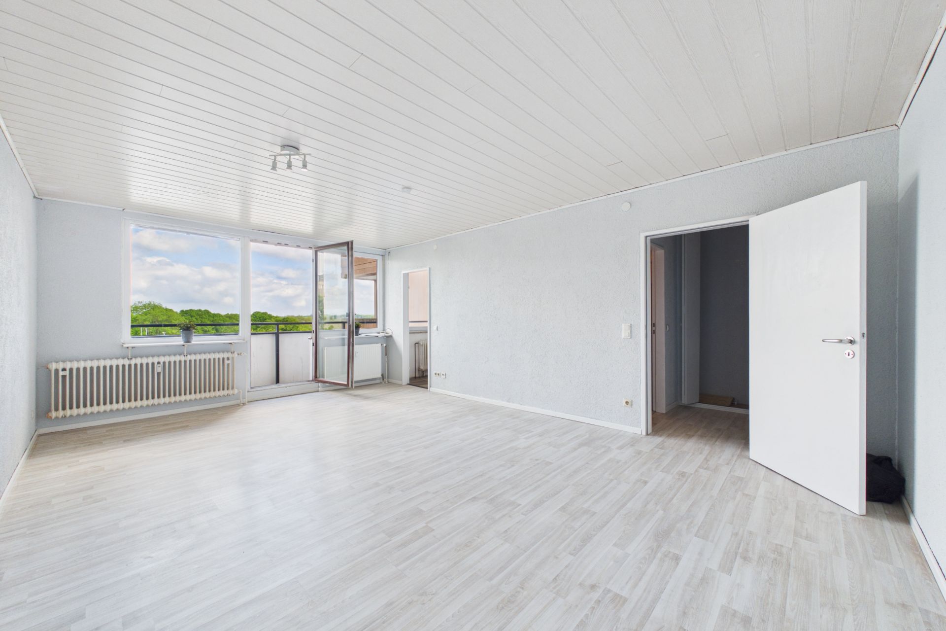 Helles, leeres Zimmer mit großen Fenstern, offenem Balkon und weißem Boden.