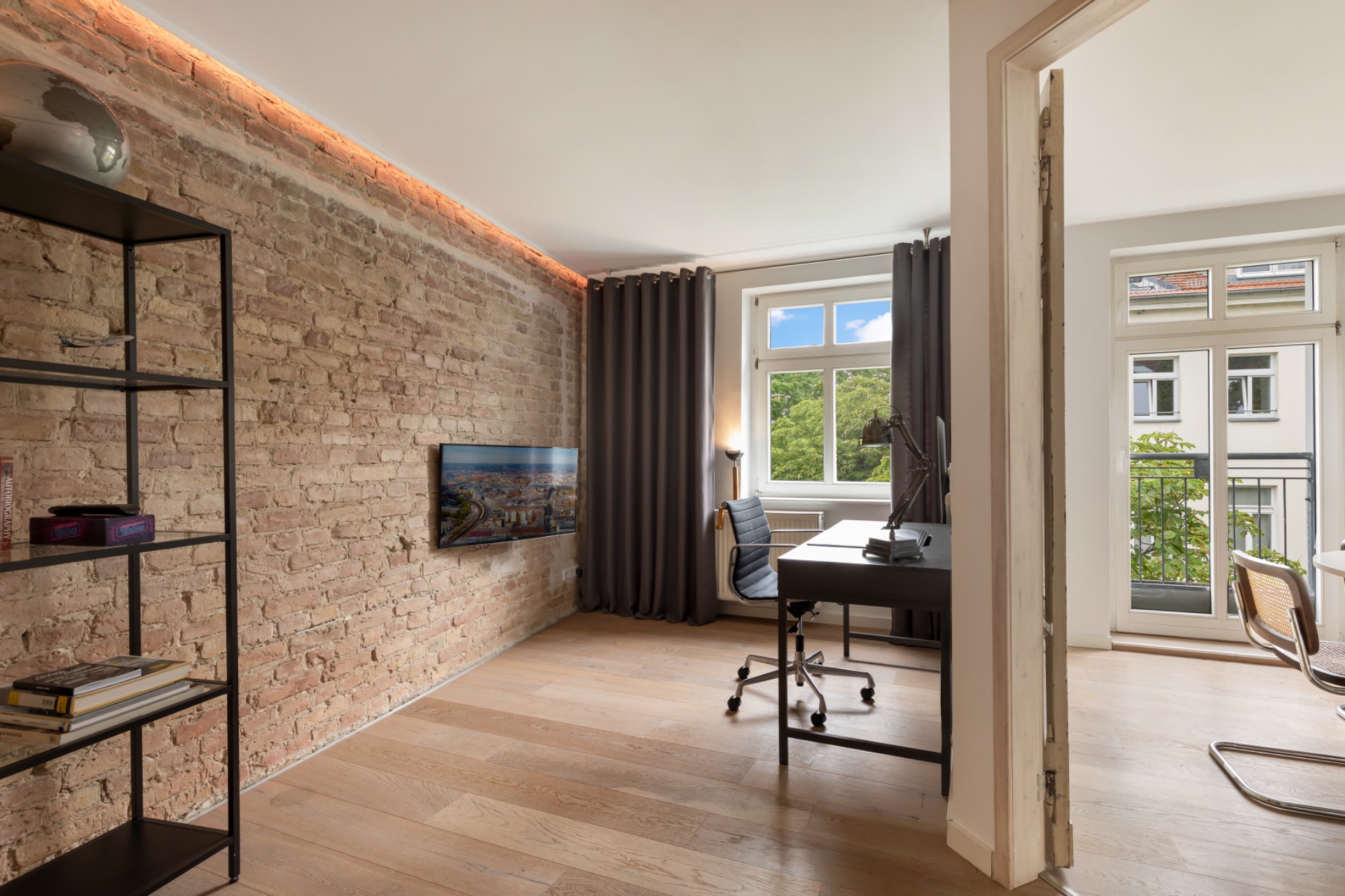 Wohnzimmer mit Backsteinwand, Schreibtisch, Stuhl, Regal, Fenster und Zugang zu hellem Raum mit Balkon.