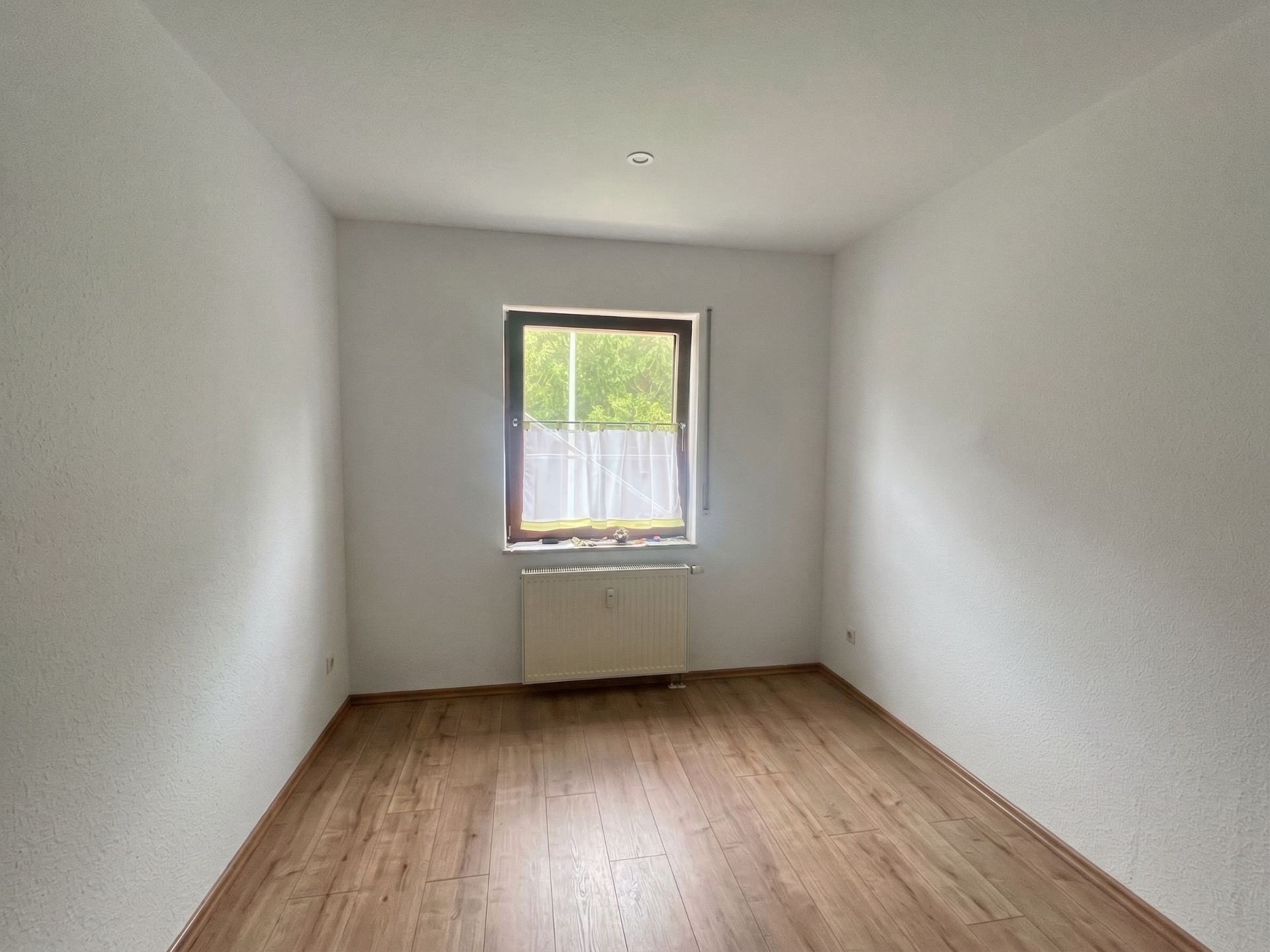 Leerer Raum mit Holzboden, weiß gestrichenen Wänden und einem Fenster mit Heizkörper darunter.