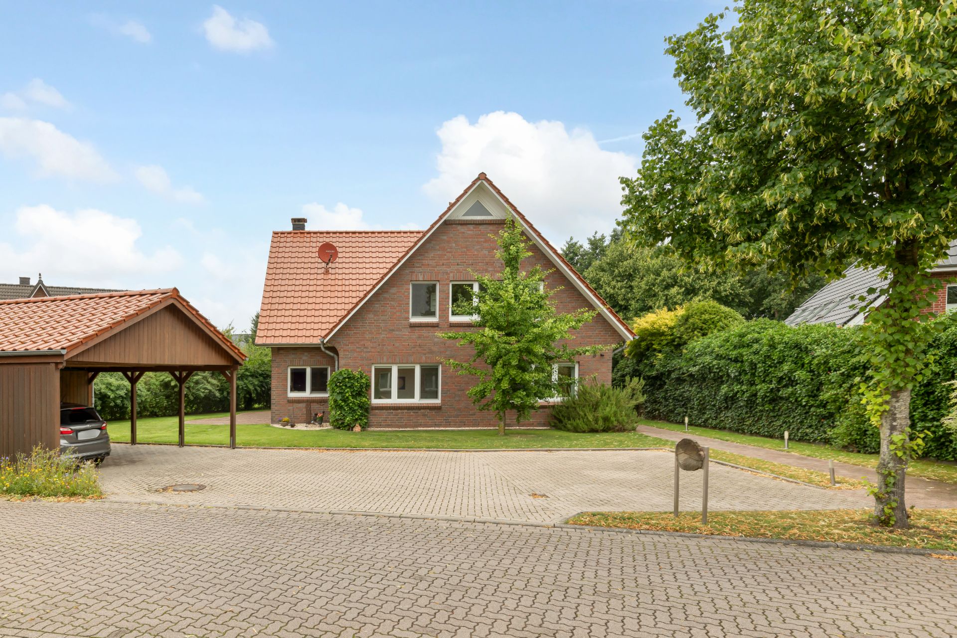 Backsteinhaus mit rotem Dach, Carport und Auto auf gepflastertem Hof, davor grüner Garten und Bäume.
