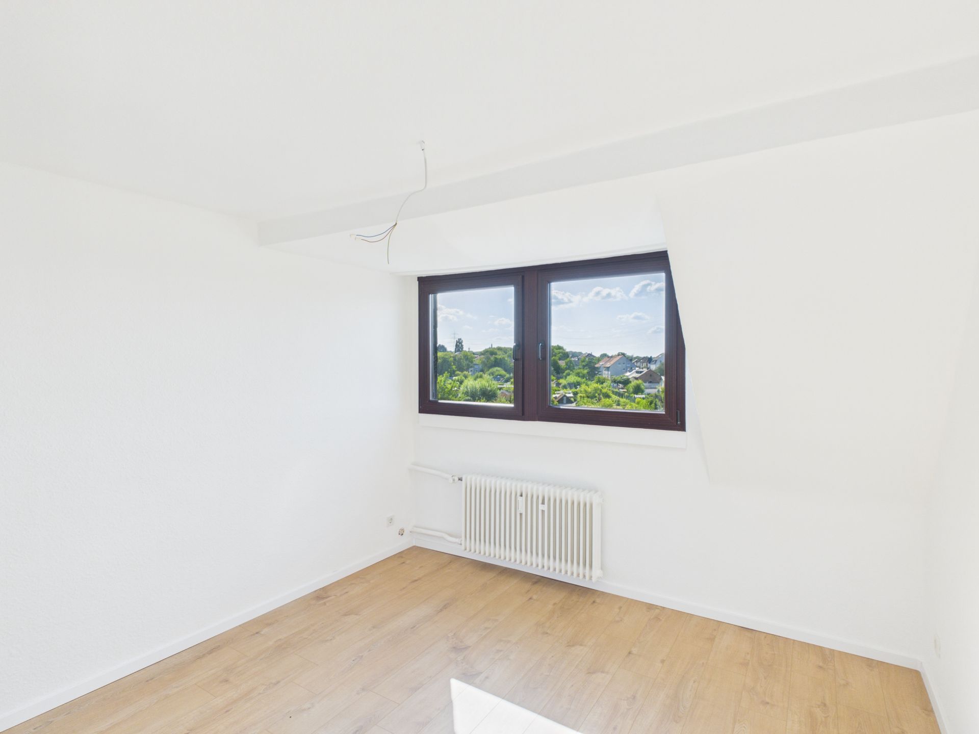 Helles, leeres Zimmer mit Holzboden, weißer Wand und großem Fenster mit Blick ins Grüne.