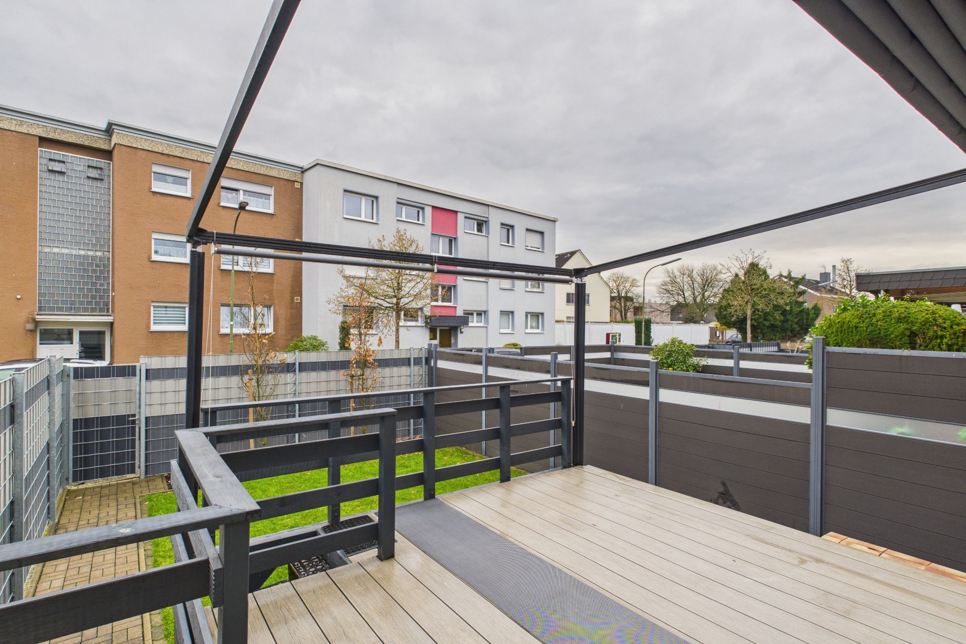 Holzterrasse mit Geländer und Blick auf einen kleinen Garten und Nachbarhäuser.