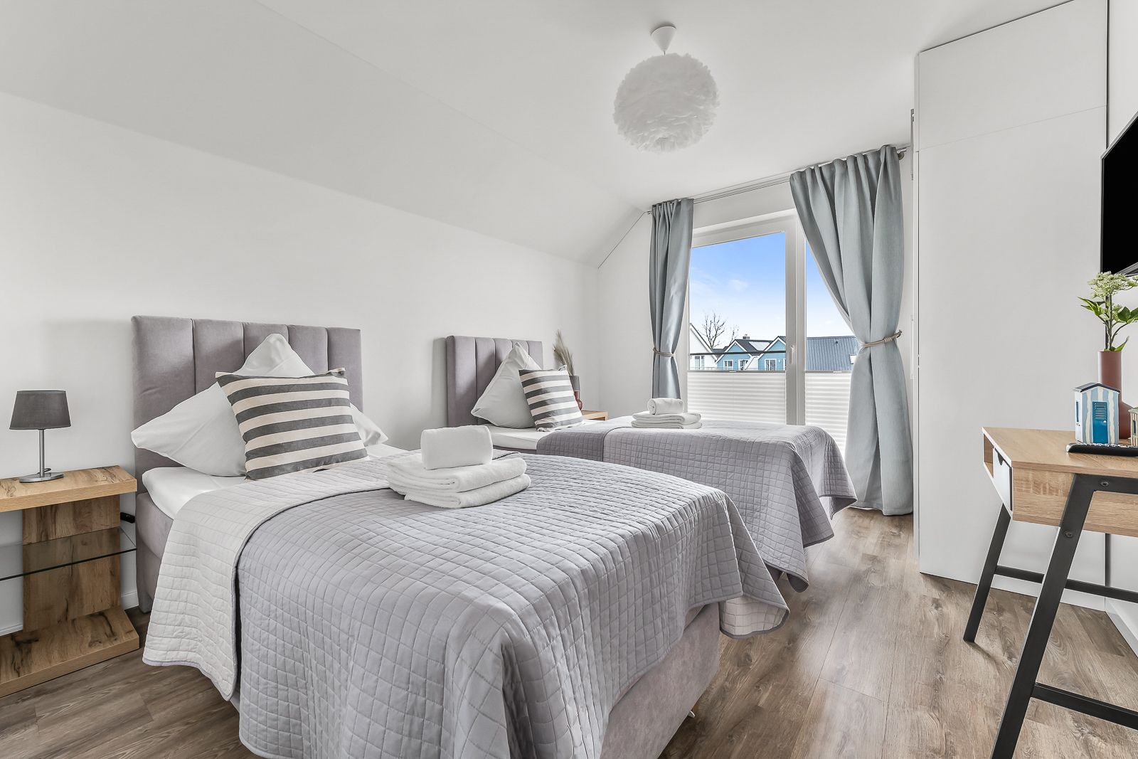 Helles Schlafzimmer mit zwei Einzelbetten, grauen Decken, Schreibtisch und großem Fenster mit Vorhängen.