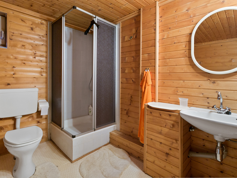 Badezimmer mit Holzwänden, Dusche, Toilette, Waschbecken und rundem Spiegel.