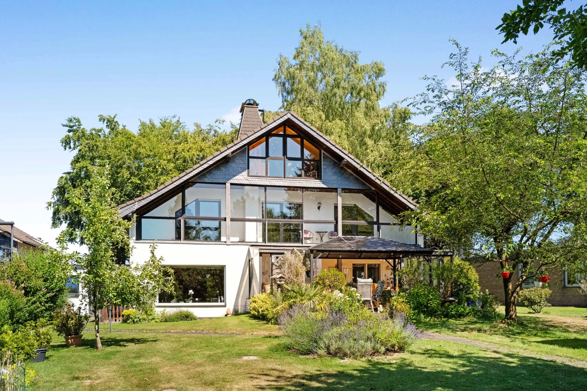 Modernes Einfamilienhaus mit großem Garten, Terrasse und vielen Fenstern, umgeben von Bäumen.
