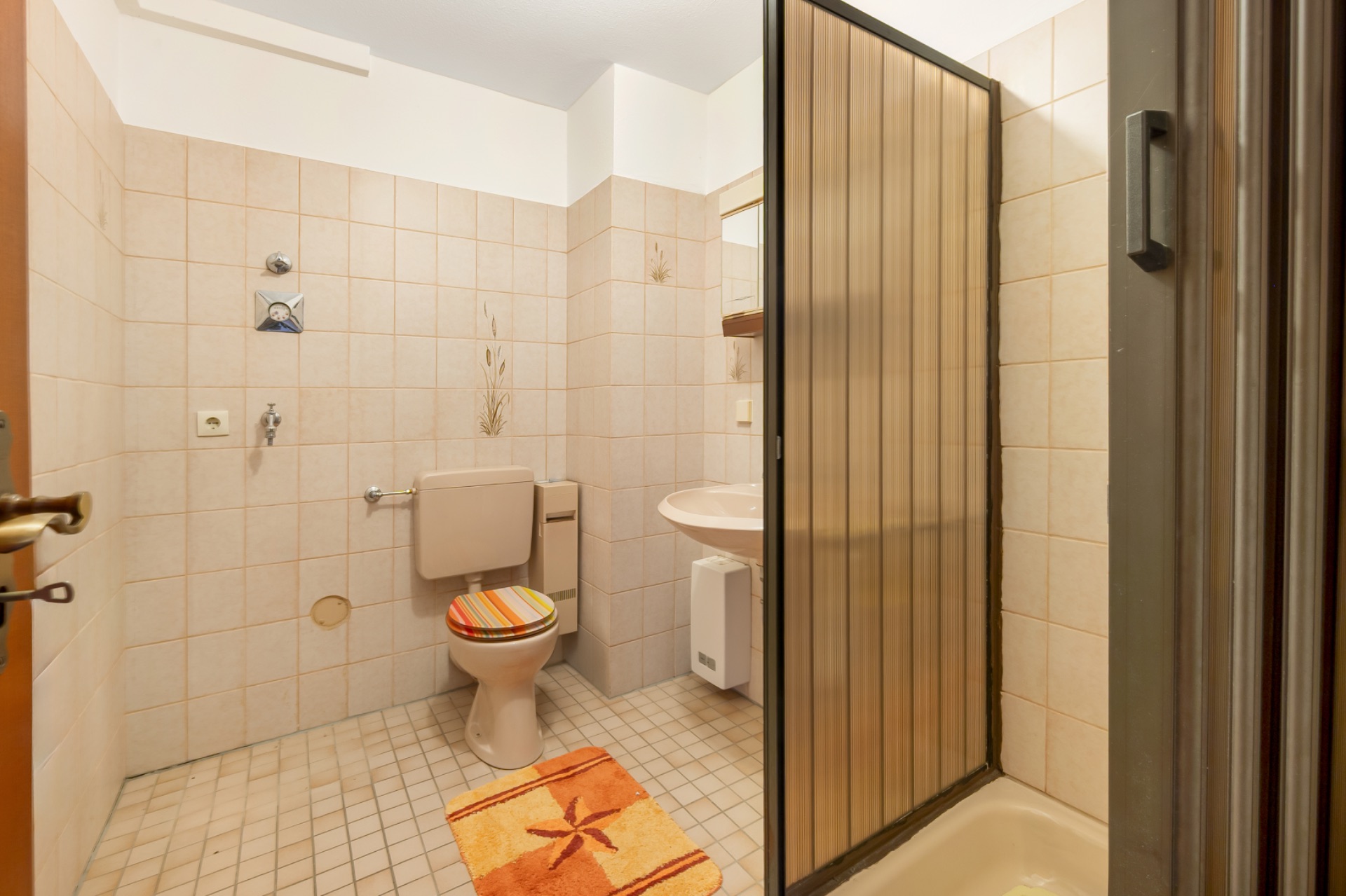 Badezimmer mit Toilette, Waschbecken und Duschkabine, orangefarbener Teppich und WC-Deckel.