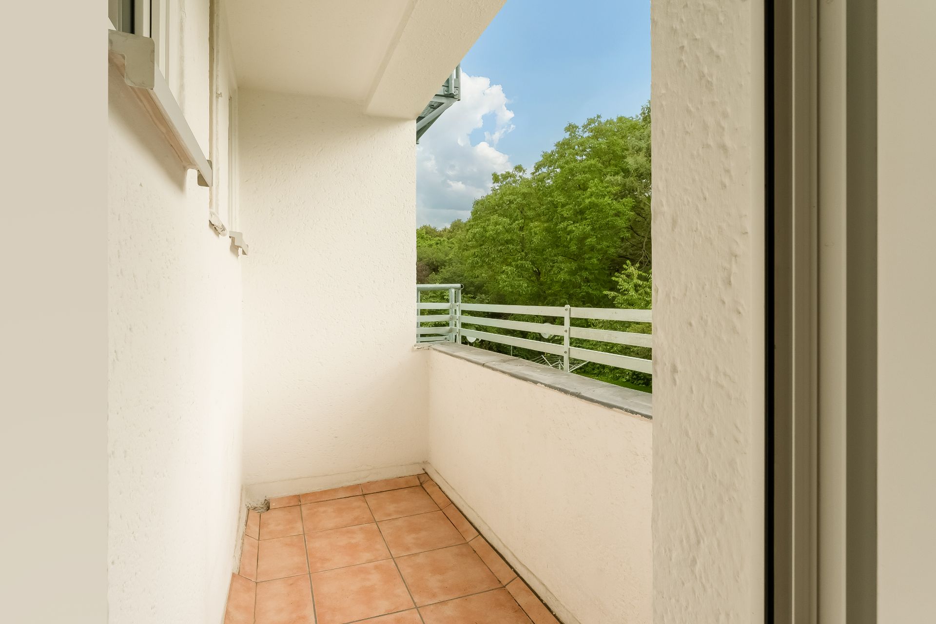 Kleiner Balkon mit Fliesenboden, weißer Wand und Blick auf grüne Bäume.