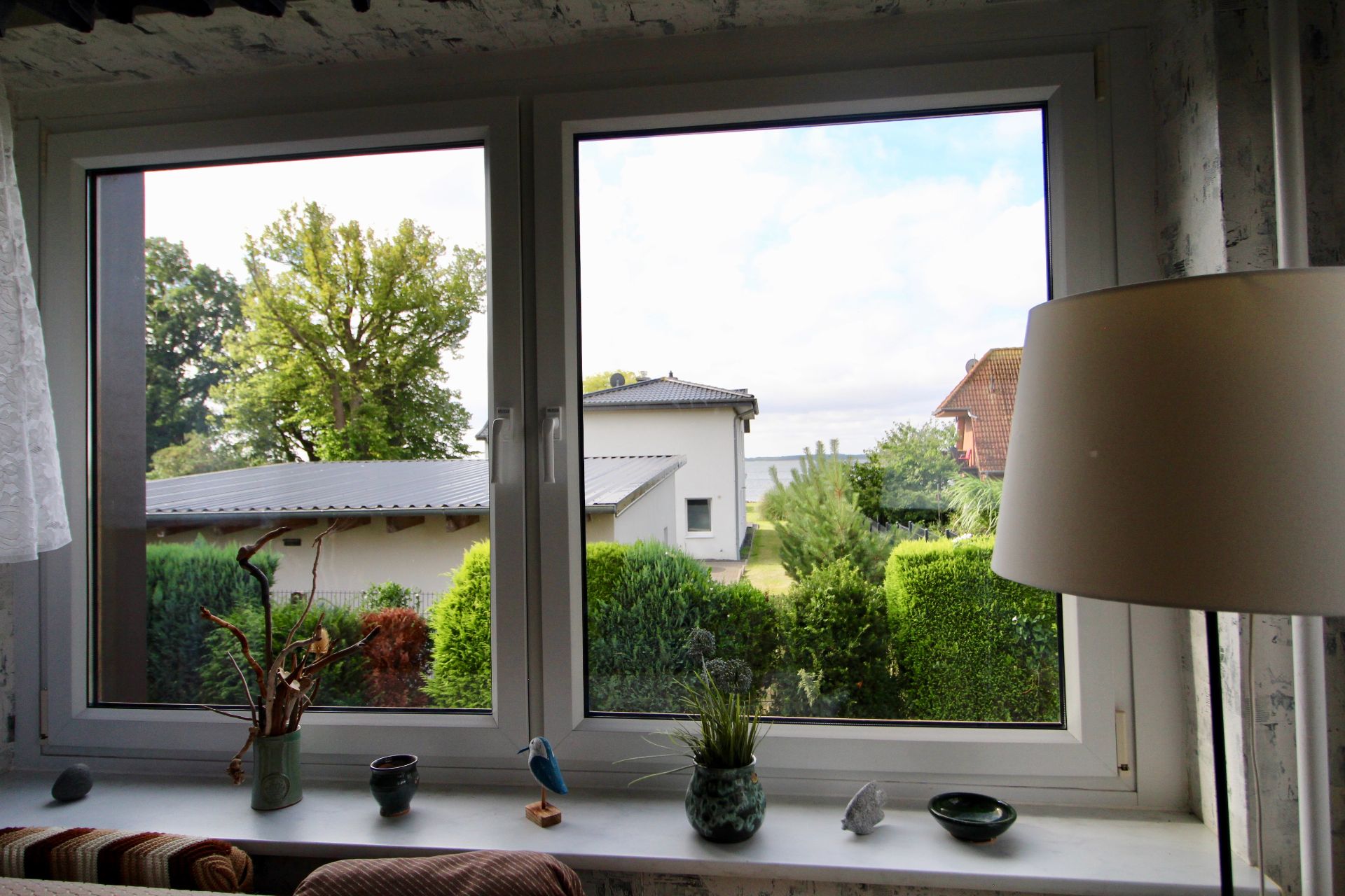 Blick durch ein großes Fenster auf Garten, Büsche und Nachbarhäuser, innen Dekorationen und eine Stehlampe.