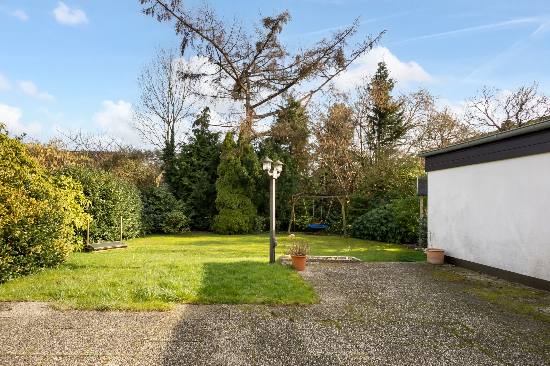 Garten mit Rasenfläche, Steinboden, Laterne, Blumentöpfen und Bäumen im Hintergrund.