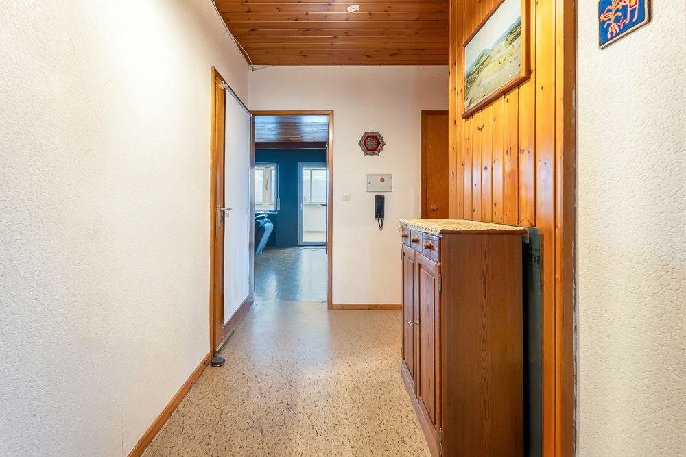 Heller Flur mit Holzmöbel, Holzdecke und Blick in ein Wohnzimmer mit großen Fenstern.