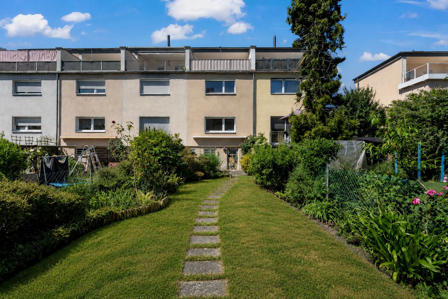 Reihenhaus mit Garten, Rasenweg führt zur Terrasse. Büsche und Pflanzen säumen den Weg.