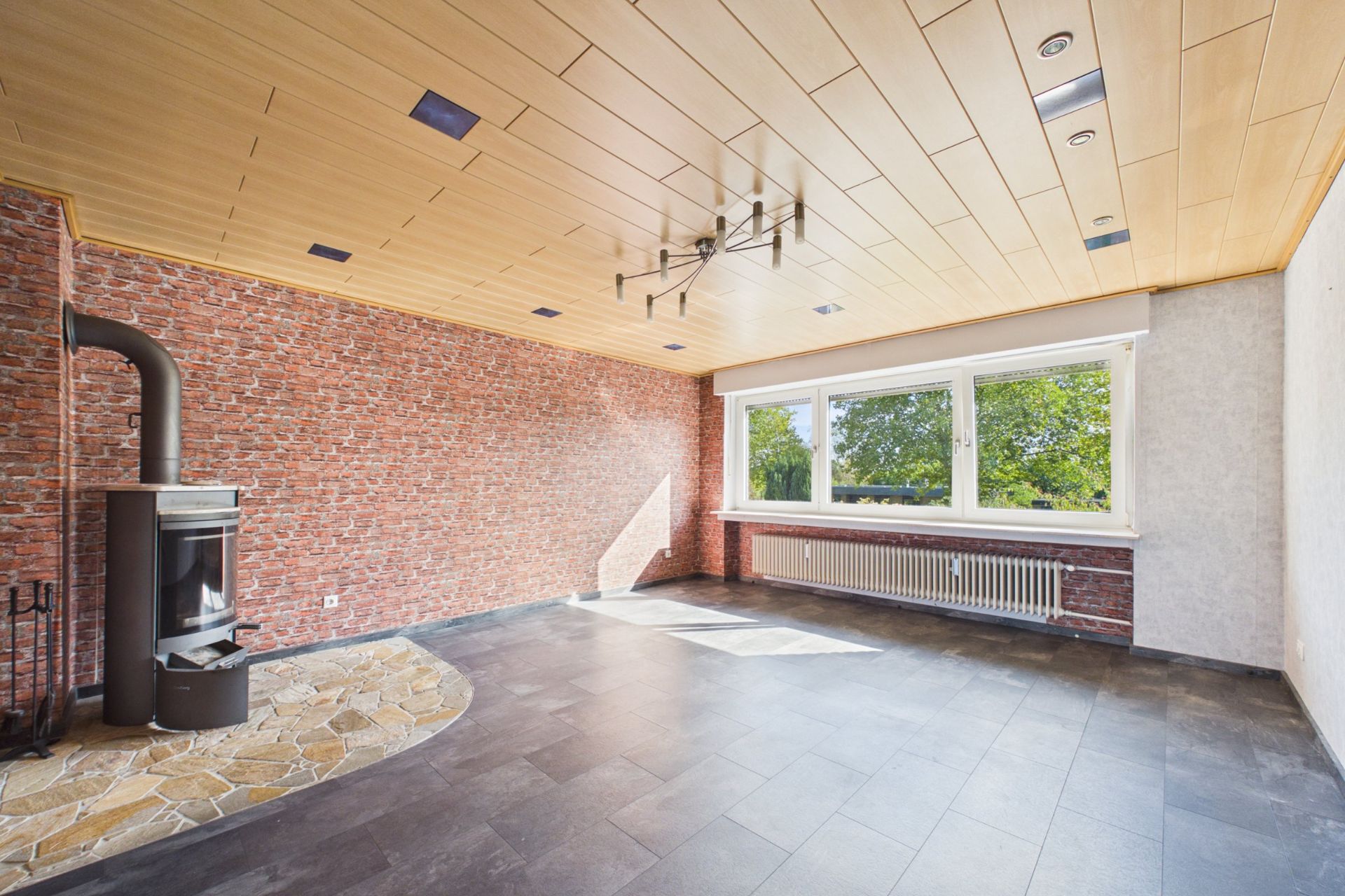 Helles Wohnzimmer mit Kaminofen, Backsteinwand und großen Fenstern mit Blick ins Grüne.
