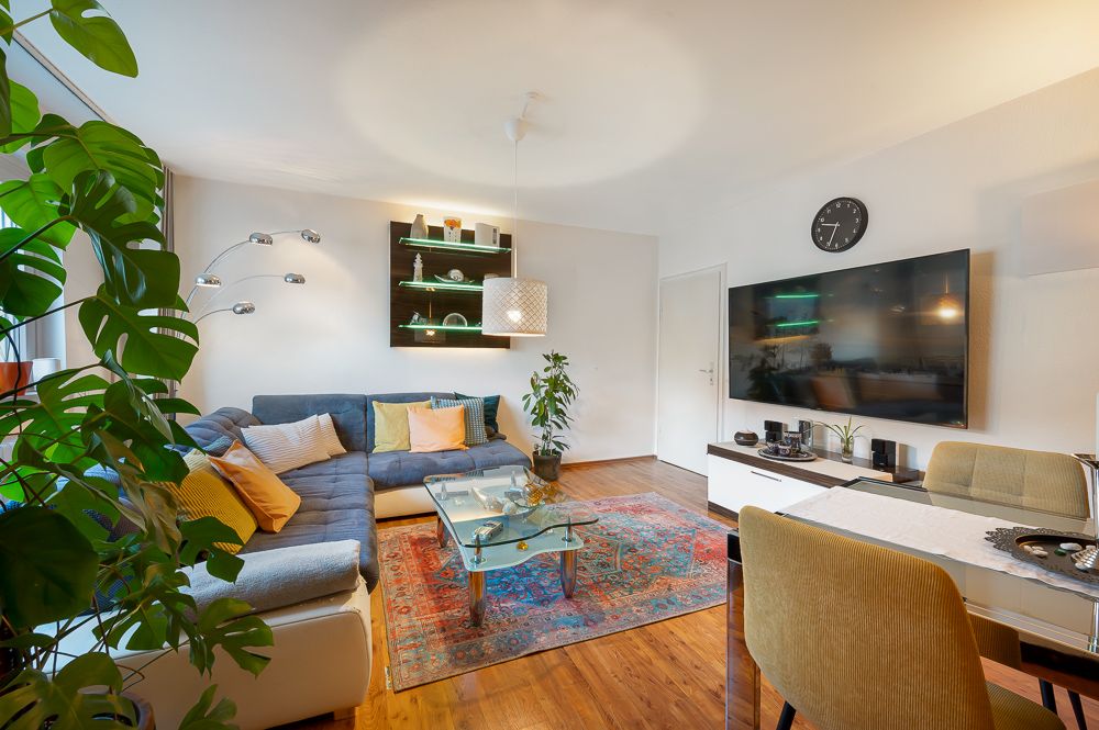 Helles Wohnzimmer mit Sofa, Teppich, Glastisch, großen Pflanzen und Wandregalen.
