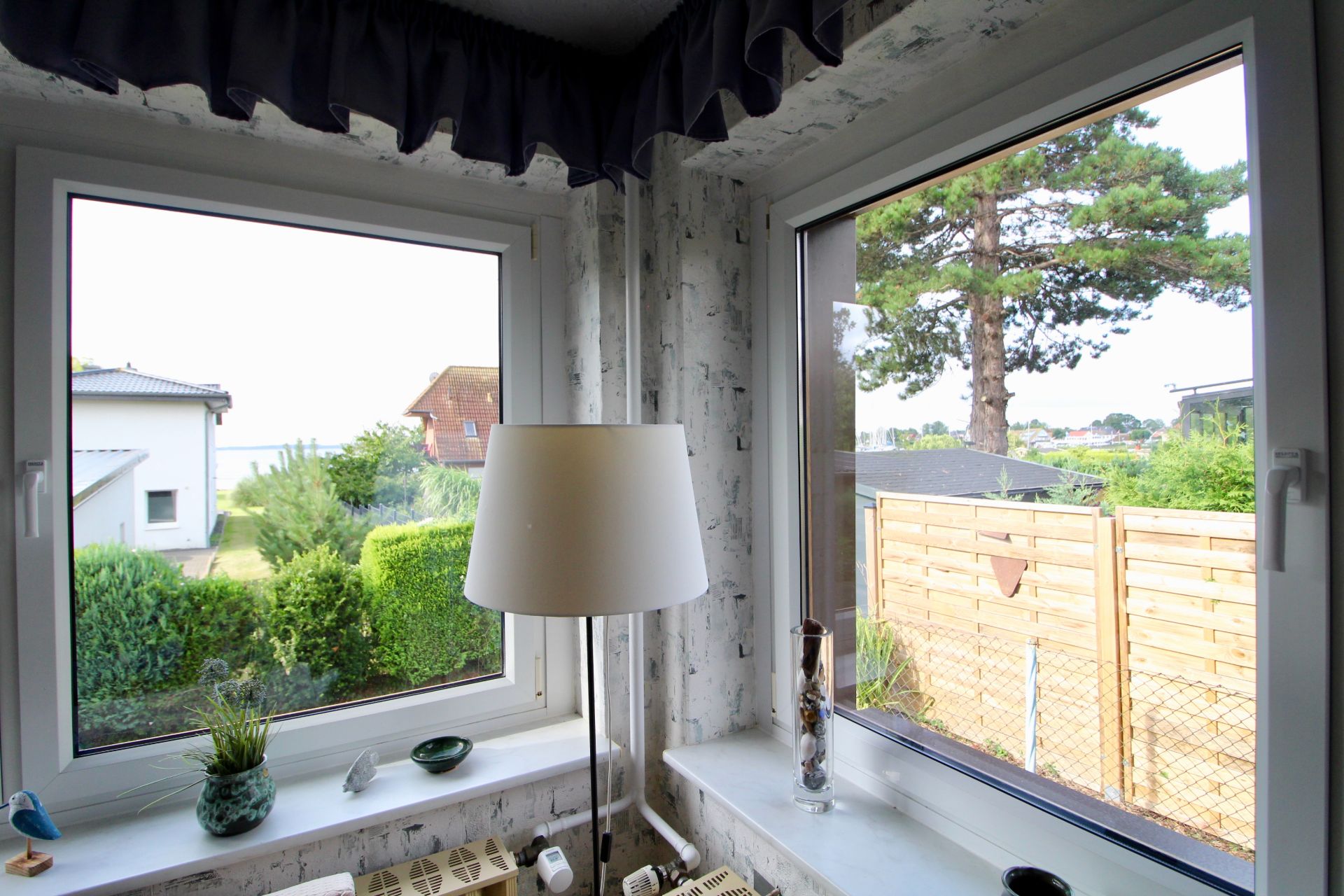 Ecke mit zwei Fenstern, Stehlampe, Pflanzen auf der Fensterbank und Blick in einen Garten.