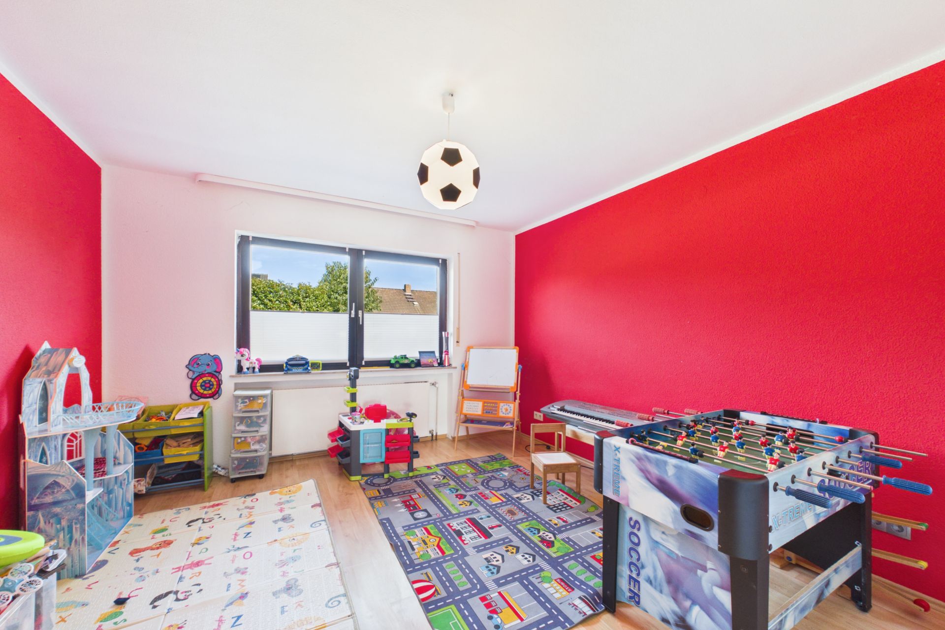 Helles Kinderzimmer mit roter Wand, Spielsachen, Malteppichen und einem Kickertisch.