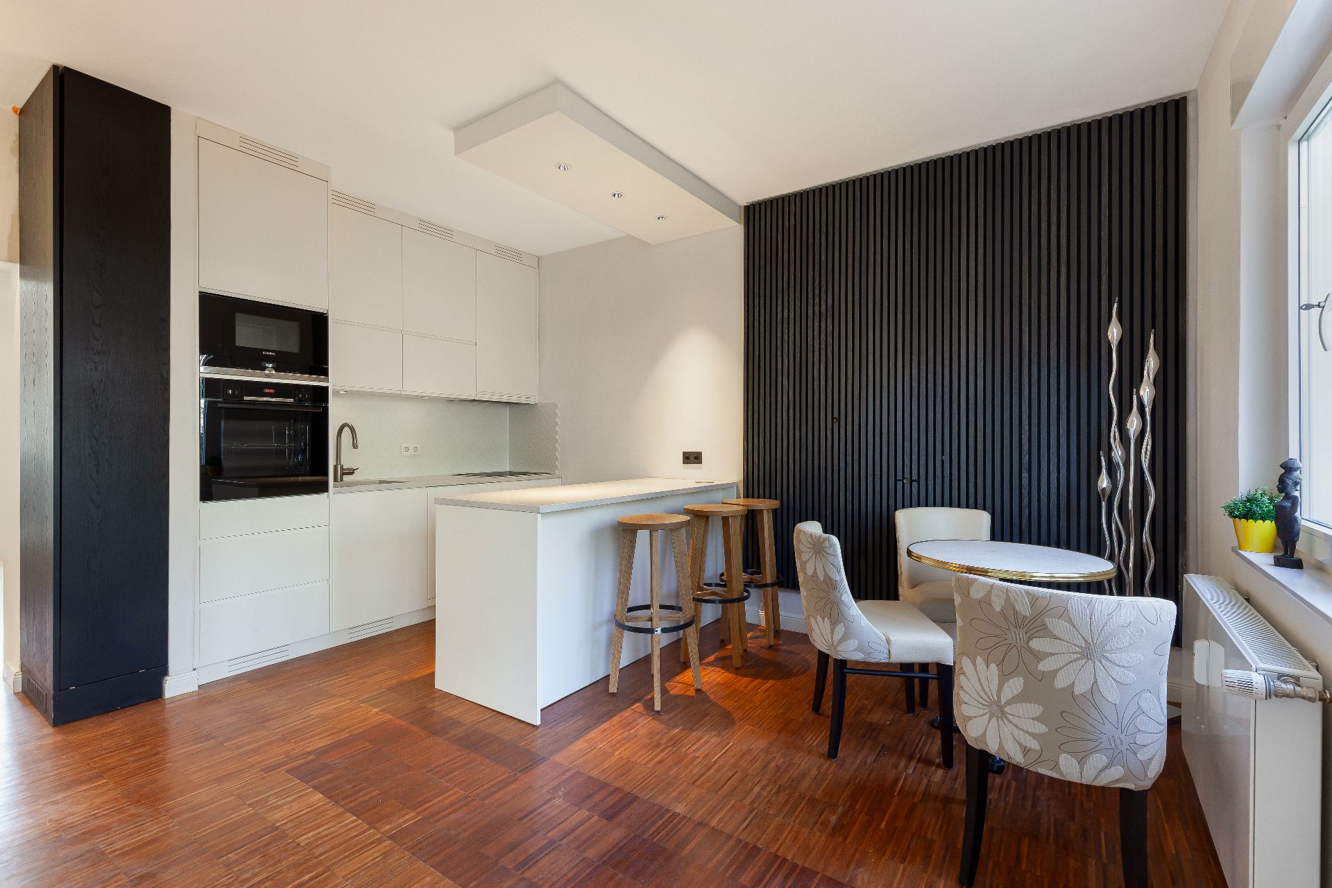 Moderne Küche mit weißer Theke, Barhockern und kleinem Esstisch mit Stühlen vor einer dunklen Wand.