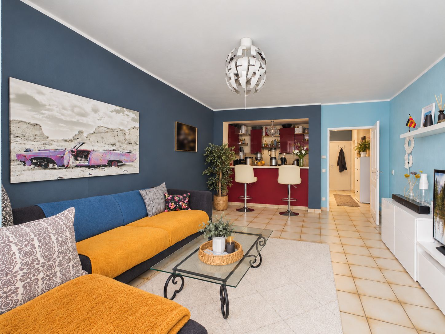Helles Wohnzimmer mit bunter Couch, Glastisch und offener Küche mit Barhockern.