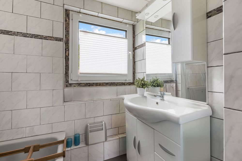 Helles Badezimmer mit weiß gekachelten Wänden, Fenster, Waschbecken, Spiegel und mehreren Pflanzen.