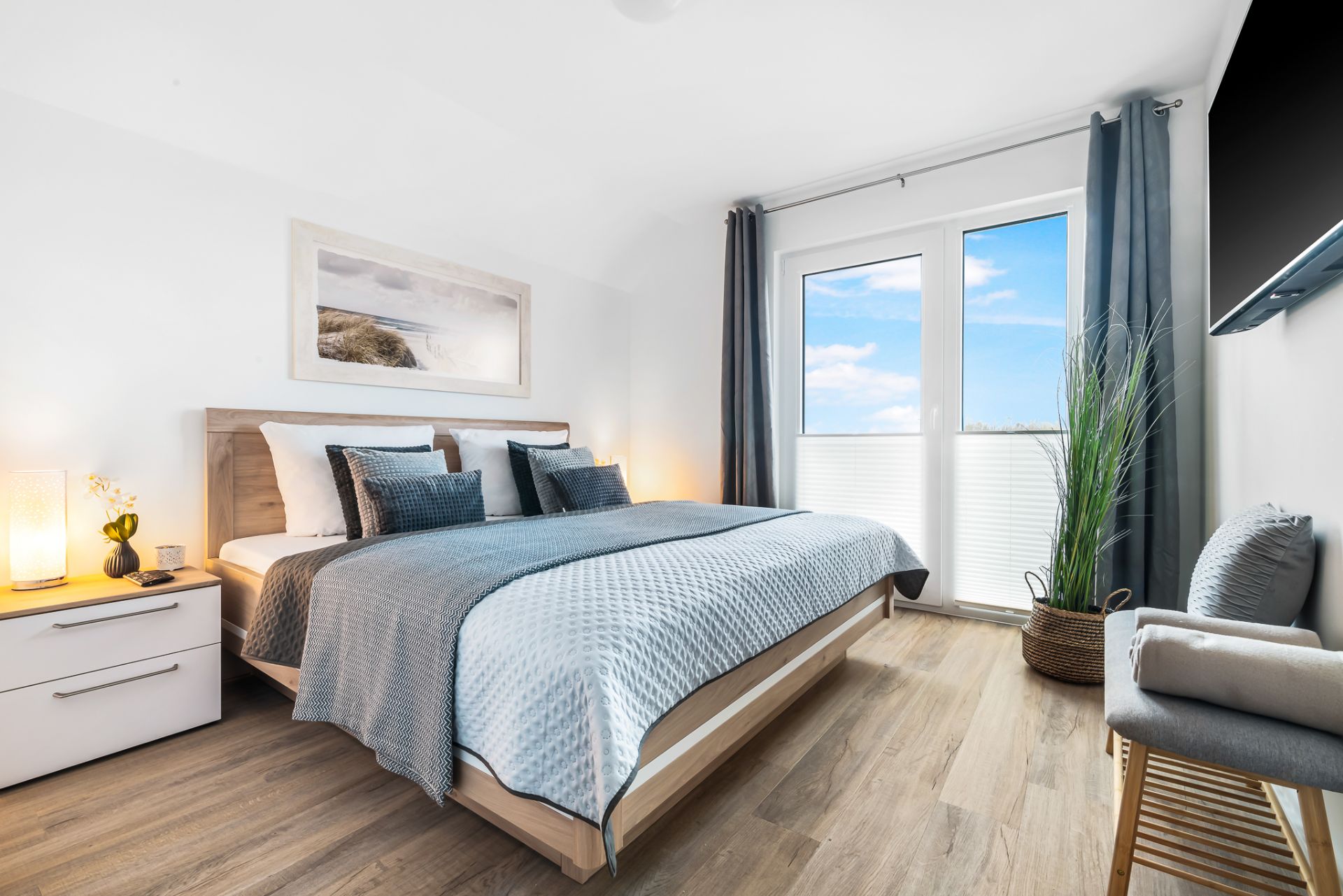 Modernes Schlafzimmer mit Doppelbett, großem Fenster, Nachttisch, Sessel und Pflanze auf Holzfußboden.