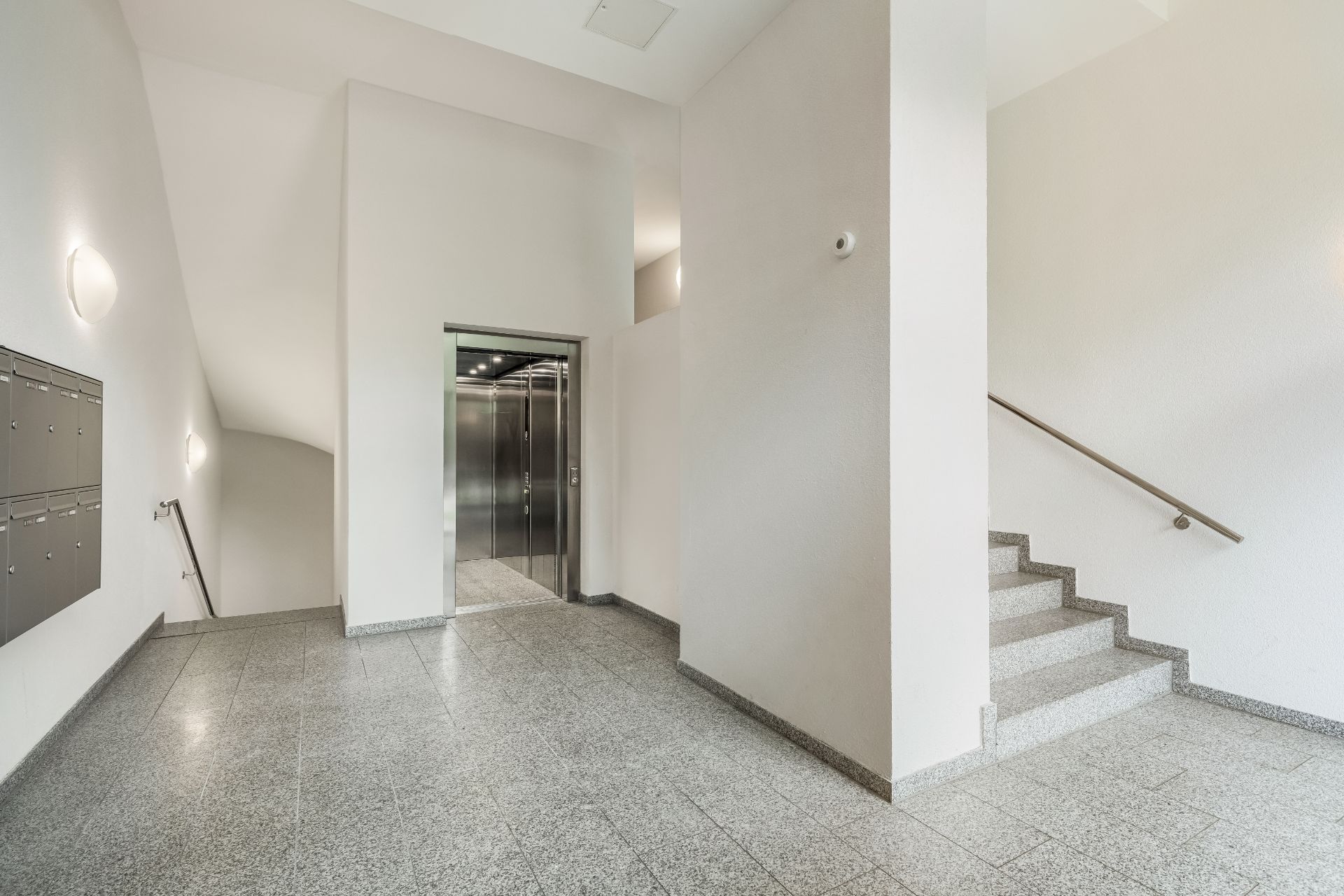 Heller Eingangsbereich mit Fahrstuhl, Treppe, Briefkästen und gefliestem Boden.