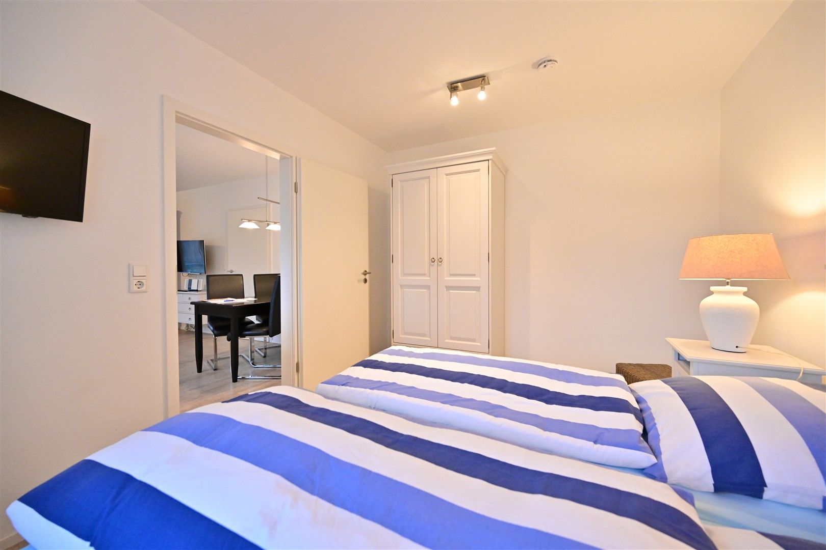 Schlafzimmer mit zwei Betten, gestreifter Bettwäsche, Kleiderschrank und Nachttisch mit Lampe.
