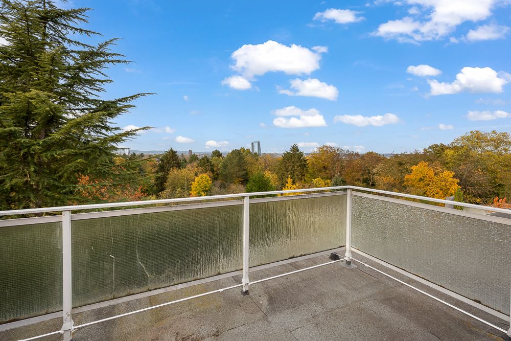 Balkon mit Glasgeländer und Blick auf Bäume und blauen Himmel mit Wolken.