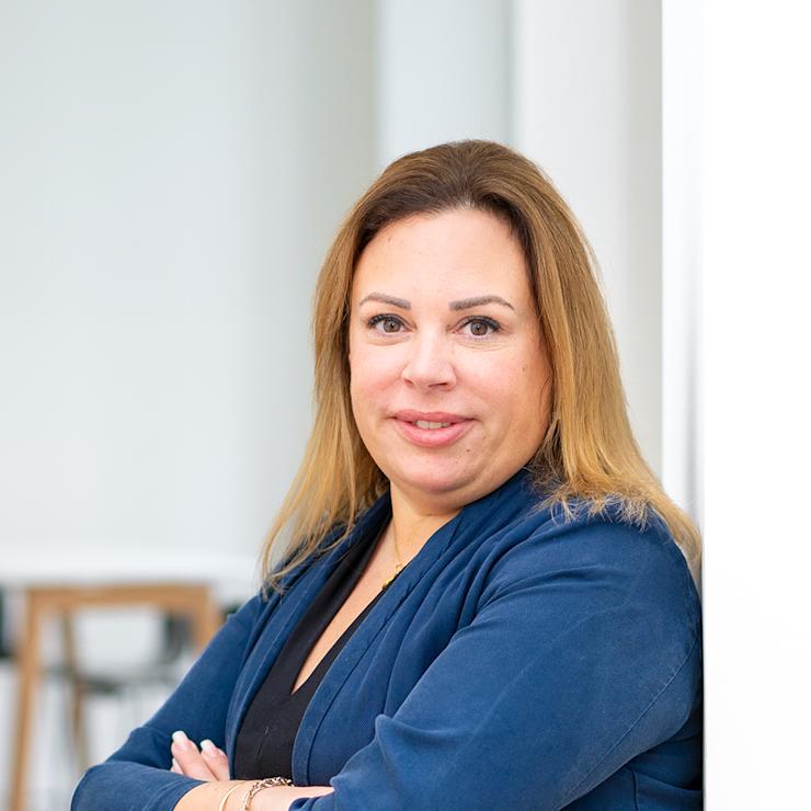 Frau mit langen Haaren und blauem Blazer steht an einer weißen Wand.