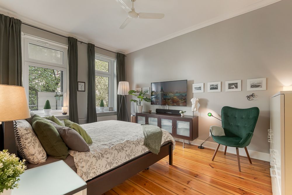 Helles Schlafzimmer mit Doppelbett, grünem Sessel, Fernseher und großen Fenstern.