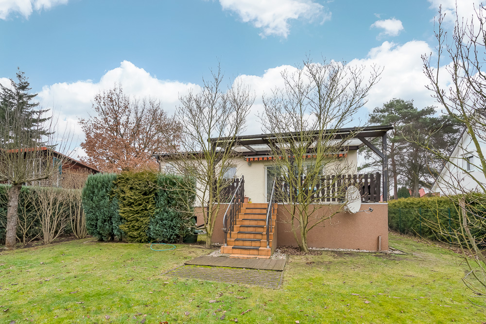 Kleines Haus mit Terrasse, Treppe und Garten, umgeben von Bäumen und Sträuchern.