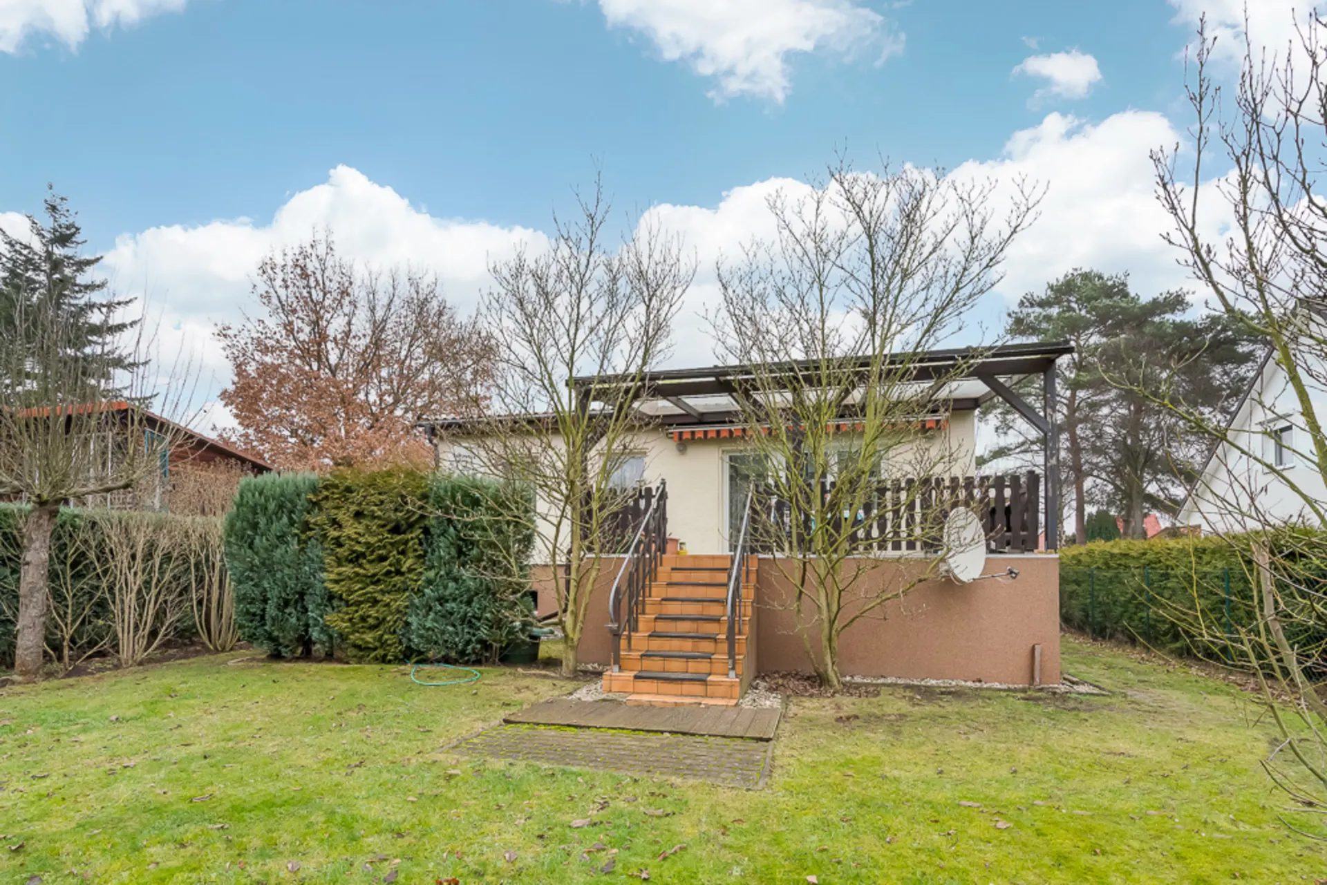 Kleines Haus mit Terrasse, Treppe und Garten, umgeben von Bäumen und Sträuchern.