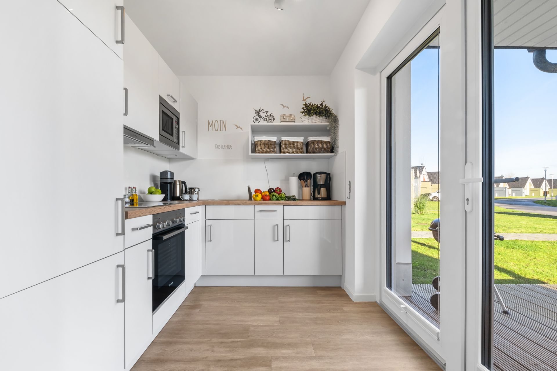 Moderne weiße Küche mit Geräten, Holzboden und großem Fenster mit Blick auf einen Garten.